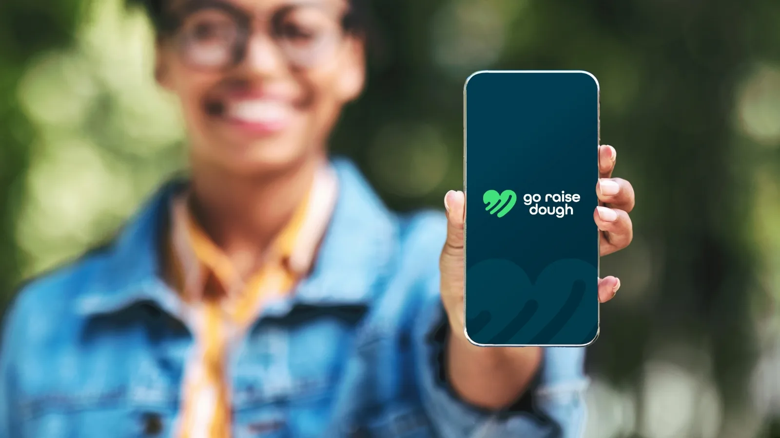 a woman holding an iPhone with go raise dough's green heart logo on a navy screen.